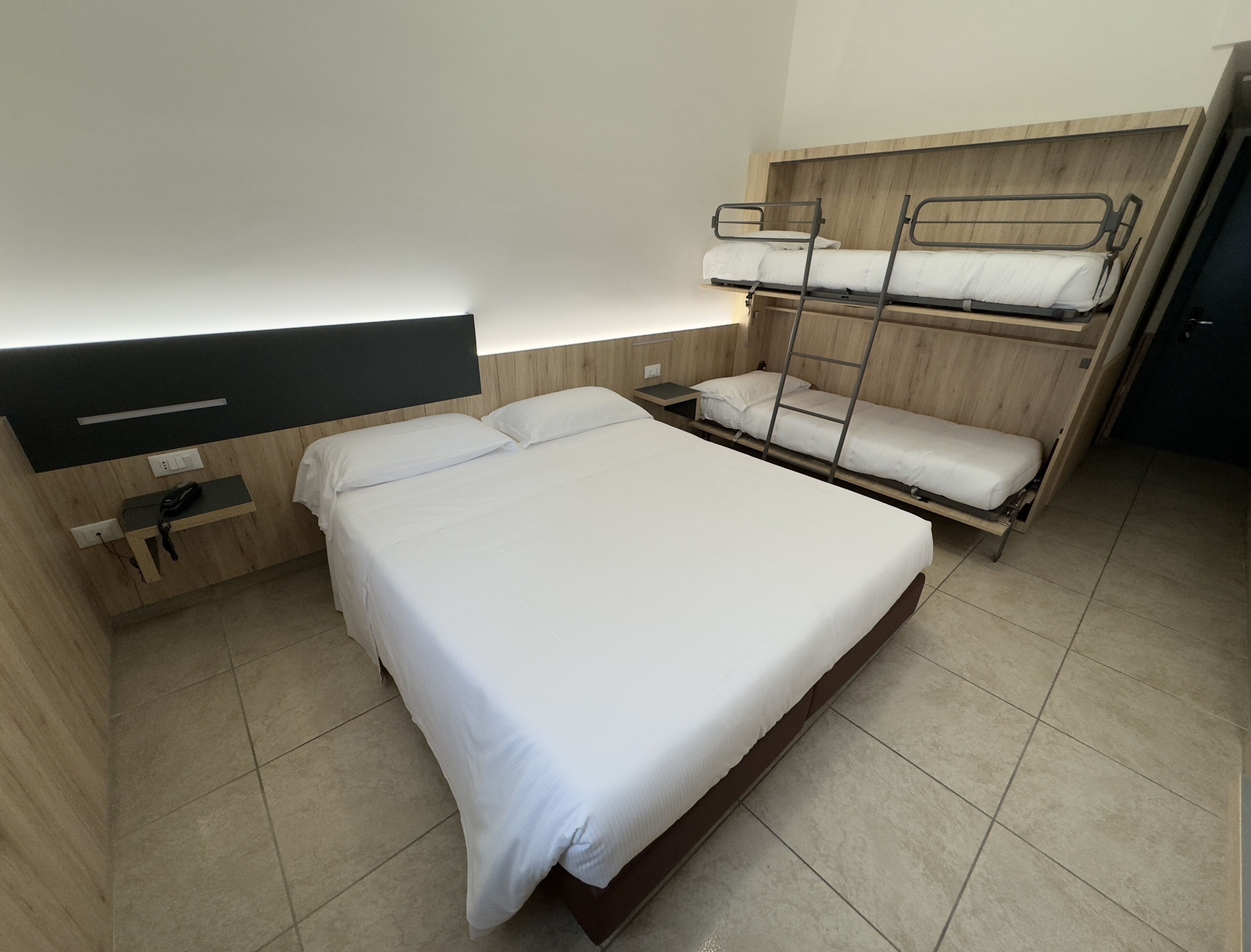 A single bed with a wooden headboard, a bedside table with a phone, and a bunk bed to the right.