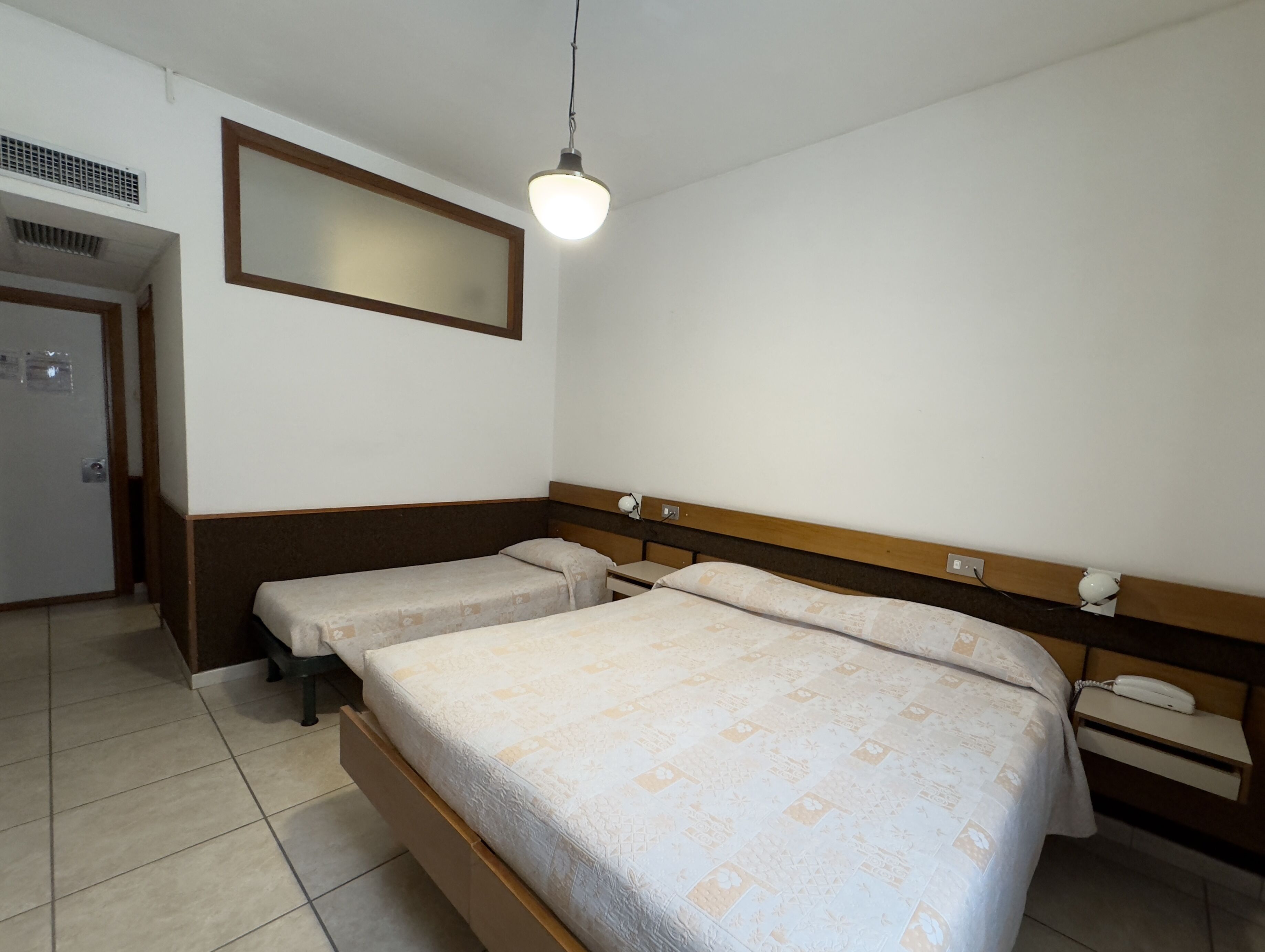 Una habitación de hotel con dos camas individuales, un cabecero de madera, una mesita de noche con lámpara y un espejo en la pared.