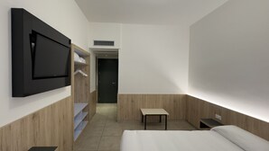Quarto de hotel moderno com cama, televisão fixada na parede, mesa pequena e guarda-roupa com prateleiras.