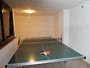 A ping pong table with a paddle in the center, a window with a dark frame, and a ceiling light.