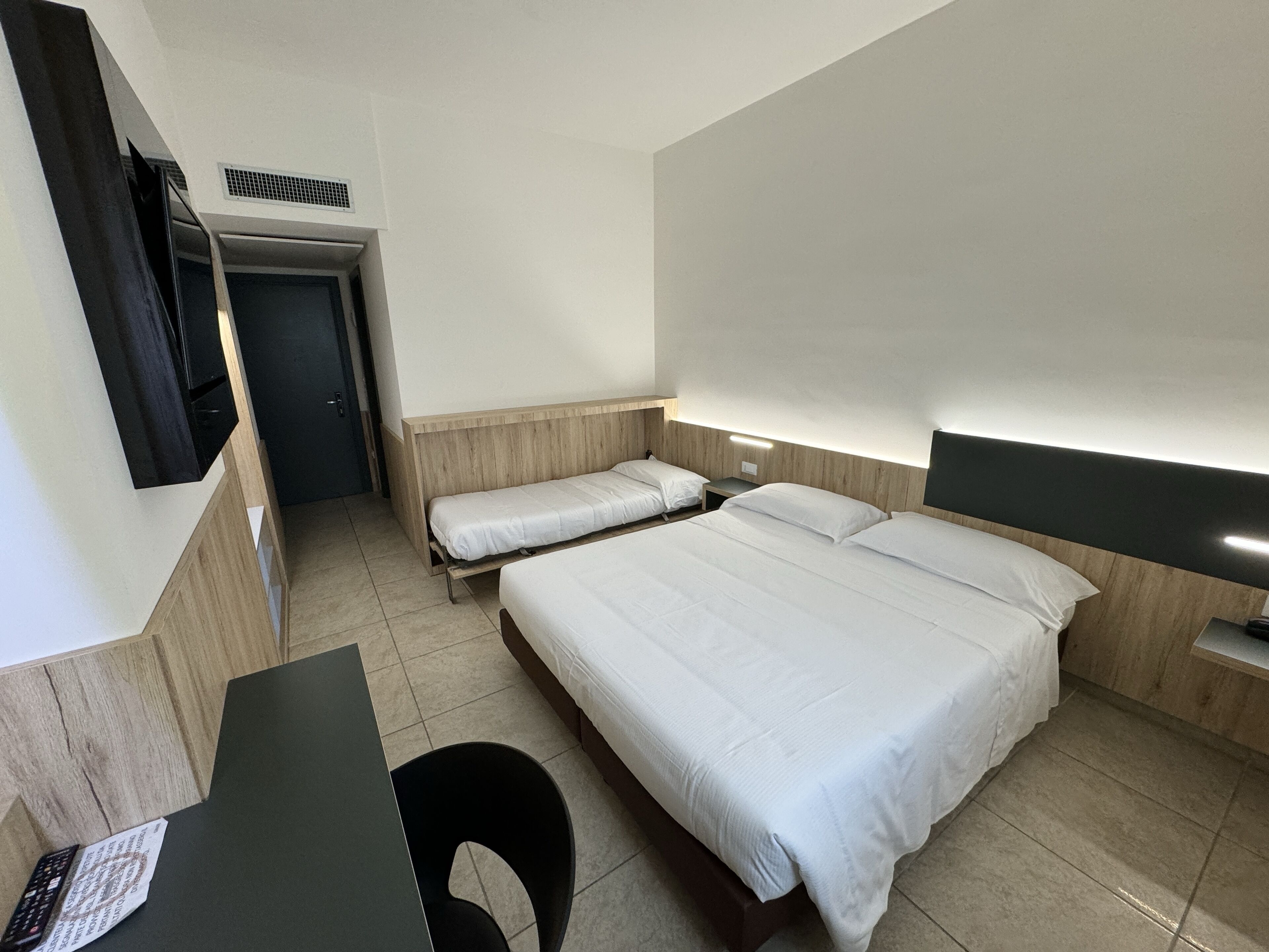 Habitación de hotel con dos camas individuales, un escritorio con silla, un televisor de pantalla plana en la pared y una mesita.