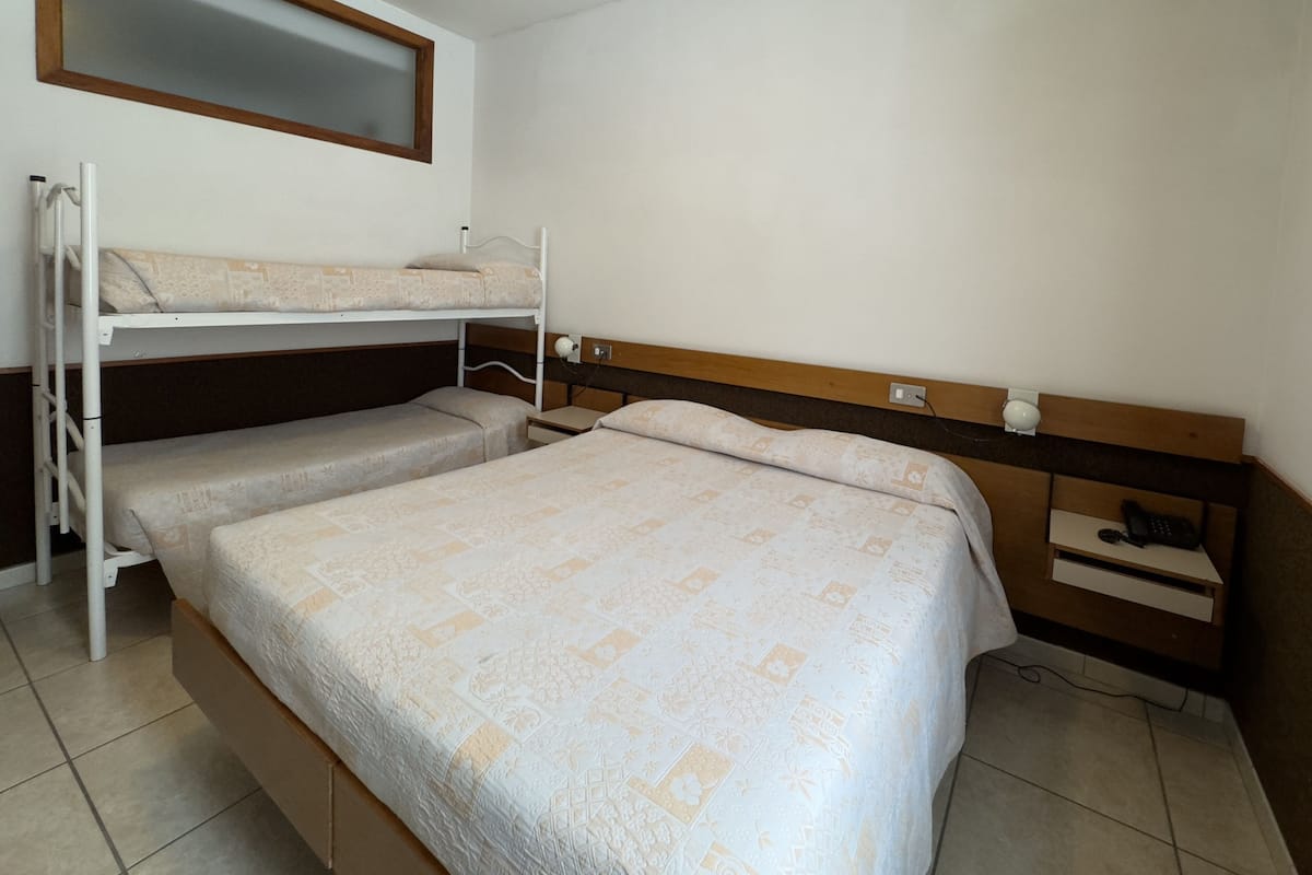 A single-bed room with a bunk bed above, a wooden headboard, a small bedside table, and a power outlet.