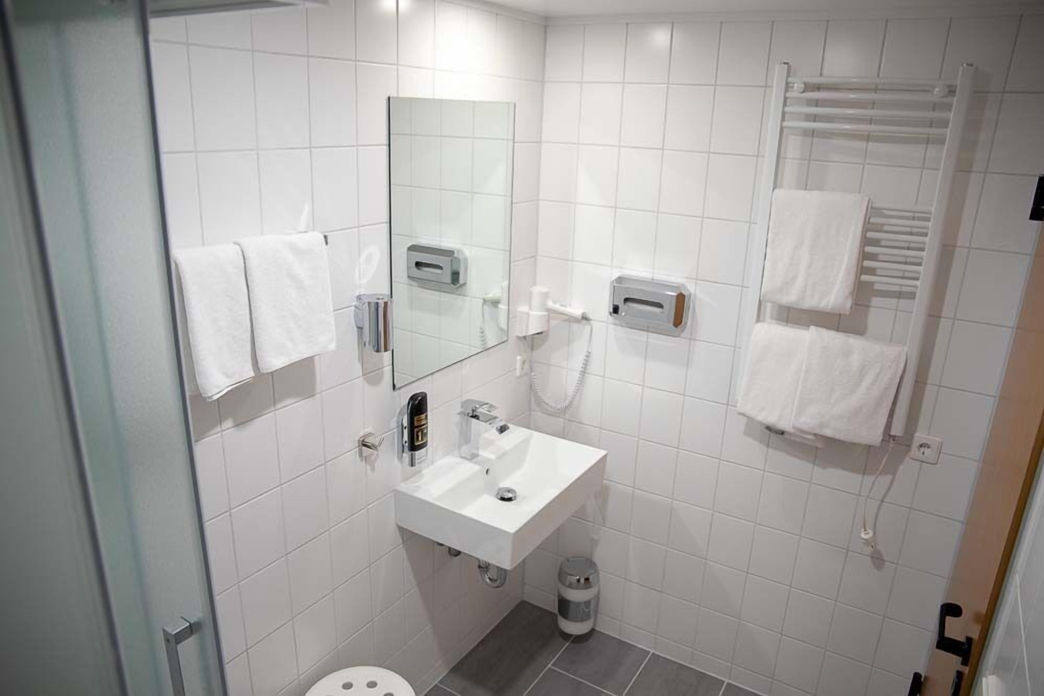Moderni kylpyhuone, jossa on valkoiset laatat, pesuallas, peili, pyyhekoukku ja roskakori.