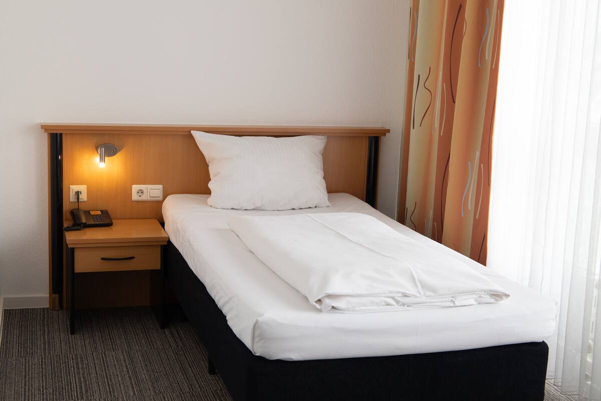 A hotel room with a neatly made bed, a bedside table with a lamp, a phone, and a wall-mounted light.