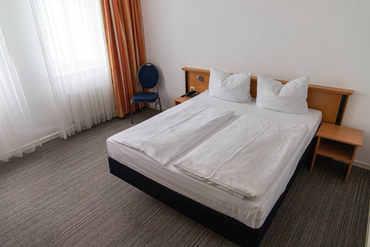 A hotel room with a large bed, a chair, a nightstand, and a window with curtains.