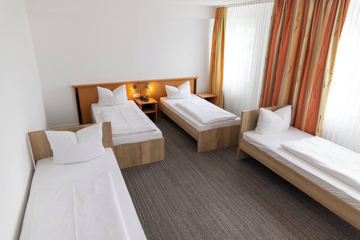 A hotel room with three single beds, a wooden headboard, a nightstand with a phone, and a window with curtains.