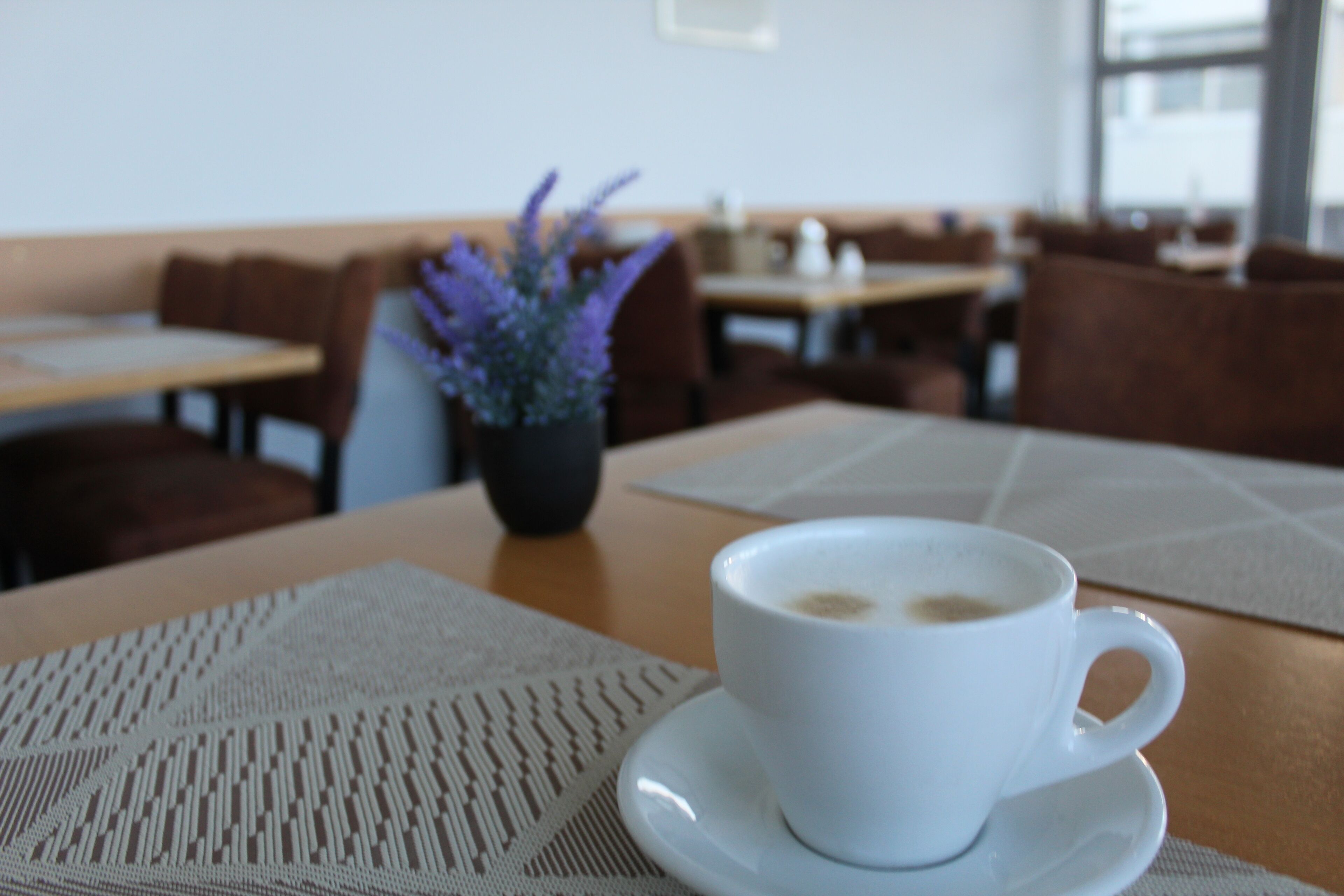 A white cup of coffee on a table with a woven placemat, a potted plant with purple flowers, and a seating area with wooden chairs and tables.