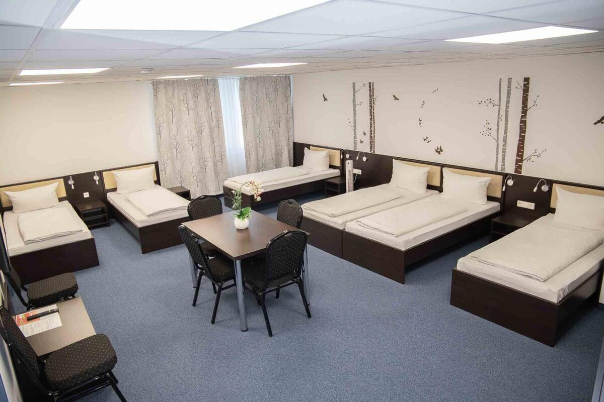A hotel room with four single beds, a small table with two chairs, and a window with curtains.