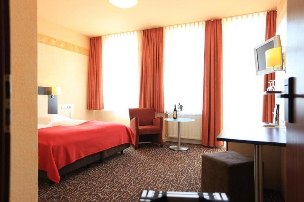 A hotel room with a bed, a chair, a small table with a bottle, and a TV on a stand.