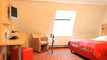 Una habitació d'hotel amb un llit, una taula de treball, una cadira, una taula petita amb una ampolla i vasos, i un televisor penjat a la paret.