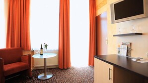 A hotel room with a red sofa, a small round table with a bottle and glasses, a TV, and a minibar. - Adesso Hotel (Kassel)