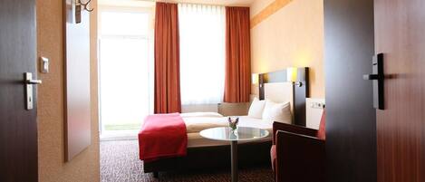 Habitación de hotel con cama, una mesita, una silla, una mesita de noche y un ventanal con cortinas.