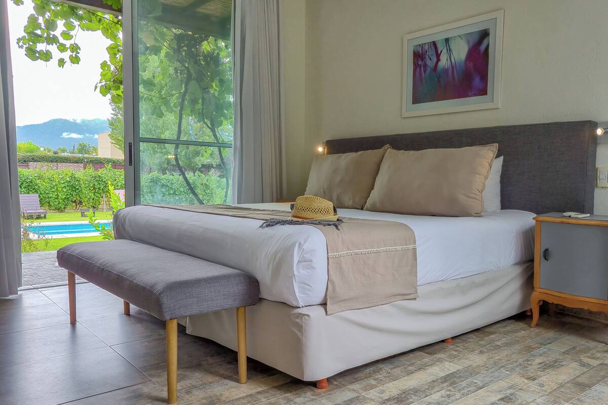 A bedroom with a bed, a bench, a nightstand, and a view of a swimming pool and greenery.