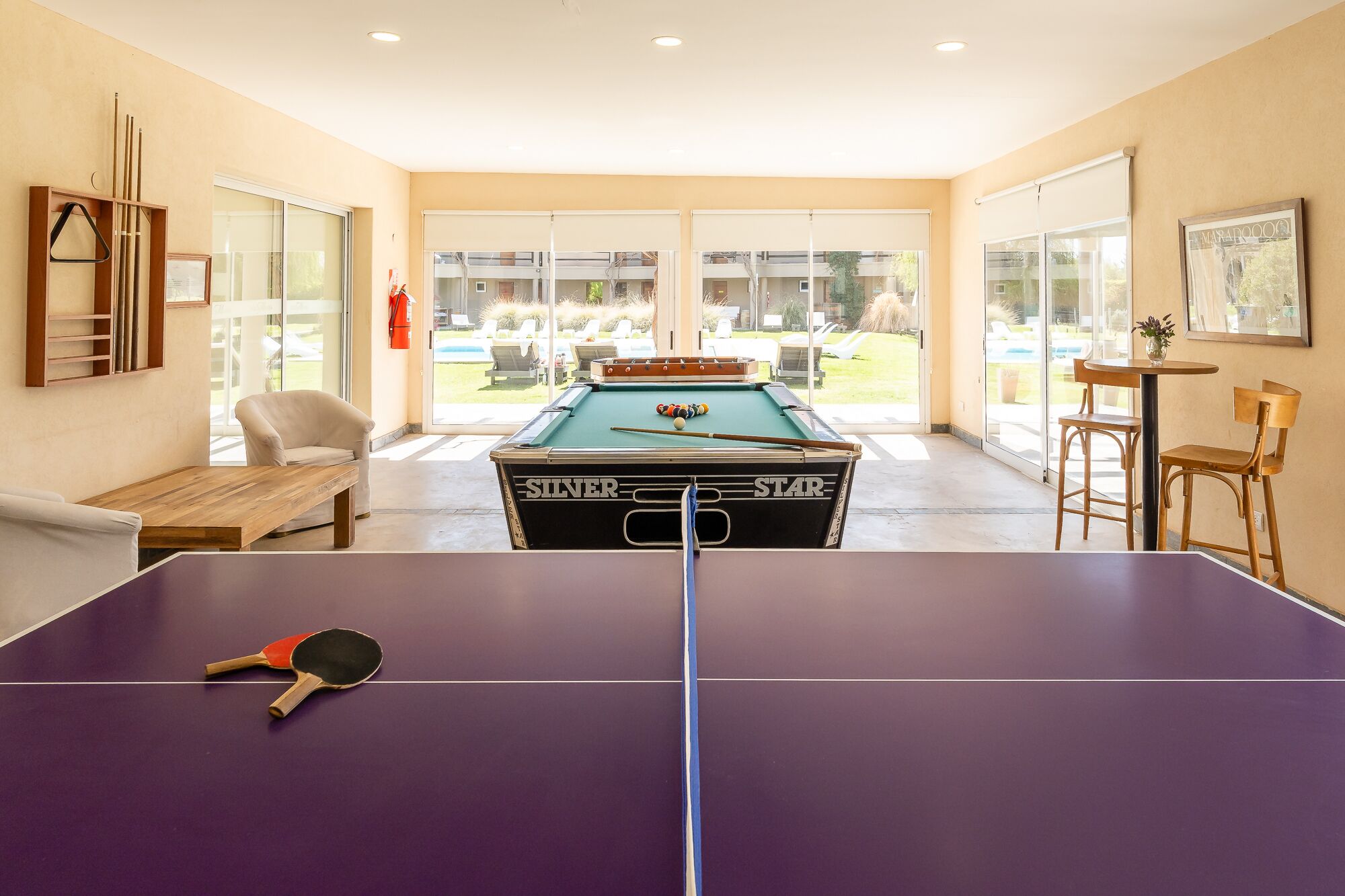 Una mesa de billar con taco y paleta de tenis de mesa, también mesa de billar y mesa de pool en una habitación con vistas a un espacio exterior.