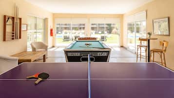 A pool table with a cue stick and a table tennis paddle, a billiards table, and a pool table in a room with a view of an outdoor area.