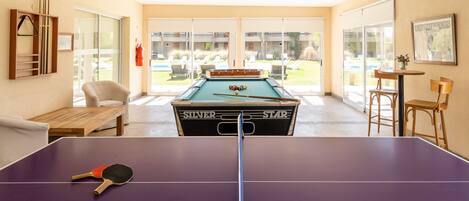 Ein Billardtisch mit Spielzeugqueue und TischtennisSchläger, ein Billardtisch und ein Pooltisch in einem Raum mit Blick auf einen Außenbereich.