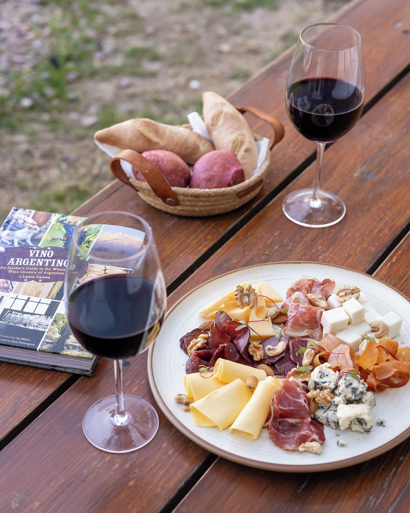 Un montaje para picnic con un libro, un plato de quesos y embutidos, y dos copas de vino tinto sobre una mesa de madera.