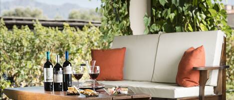 A wooden table with wine bottles, glasses, and food items on a patio with a white cushioned bench and orange pillows.