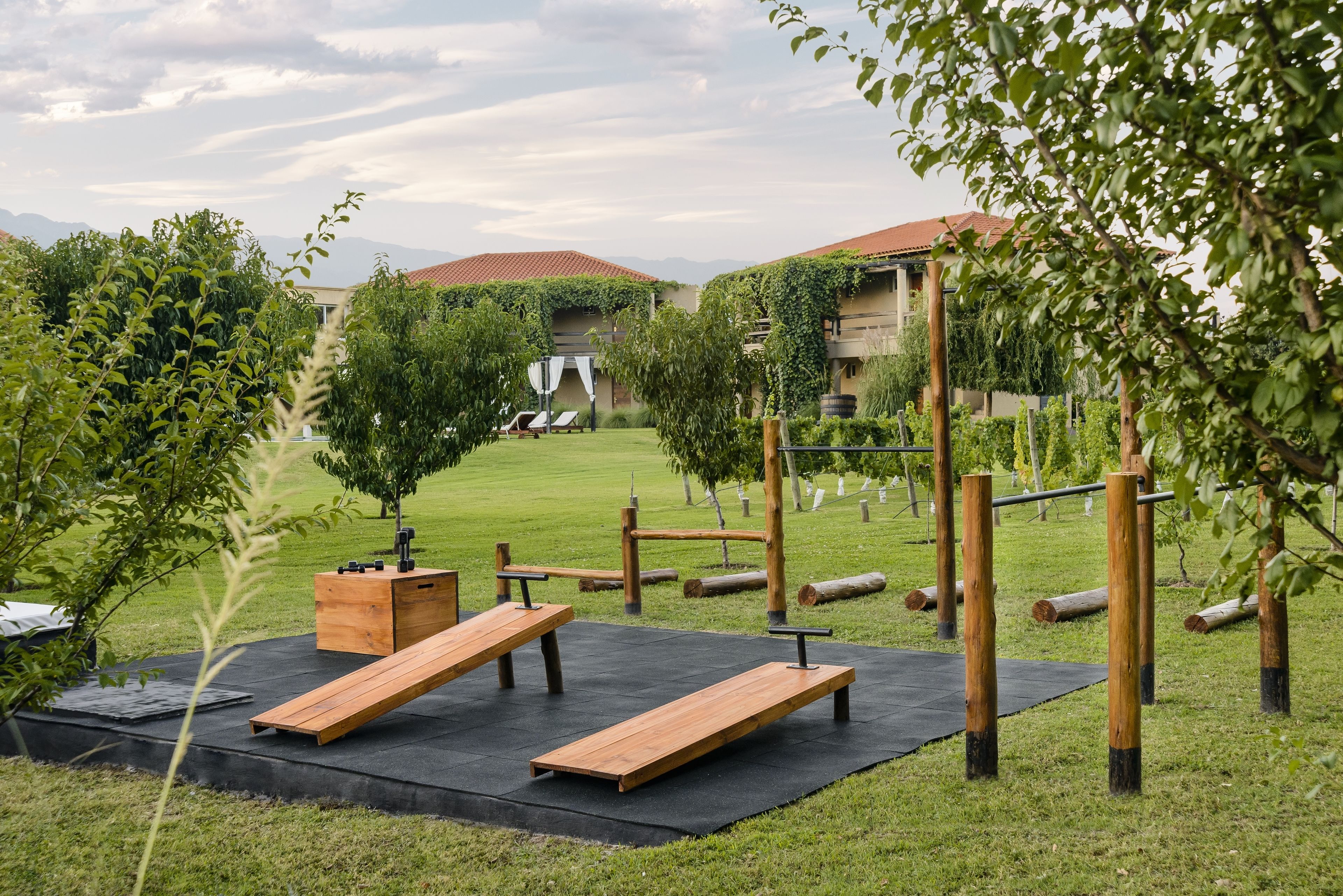 Zona de gimnasio al aire libre con bancos de madera y equipo de ejercicio, rodeada de follaje y un edificio con techo de tejas rojas al fondo.