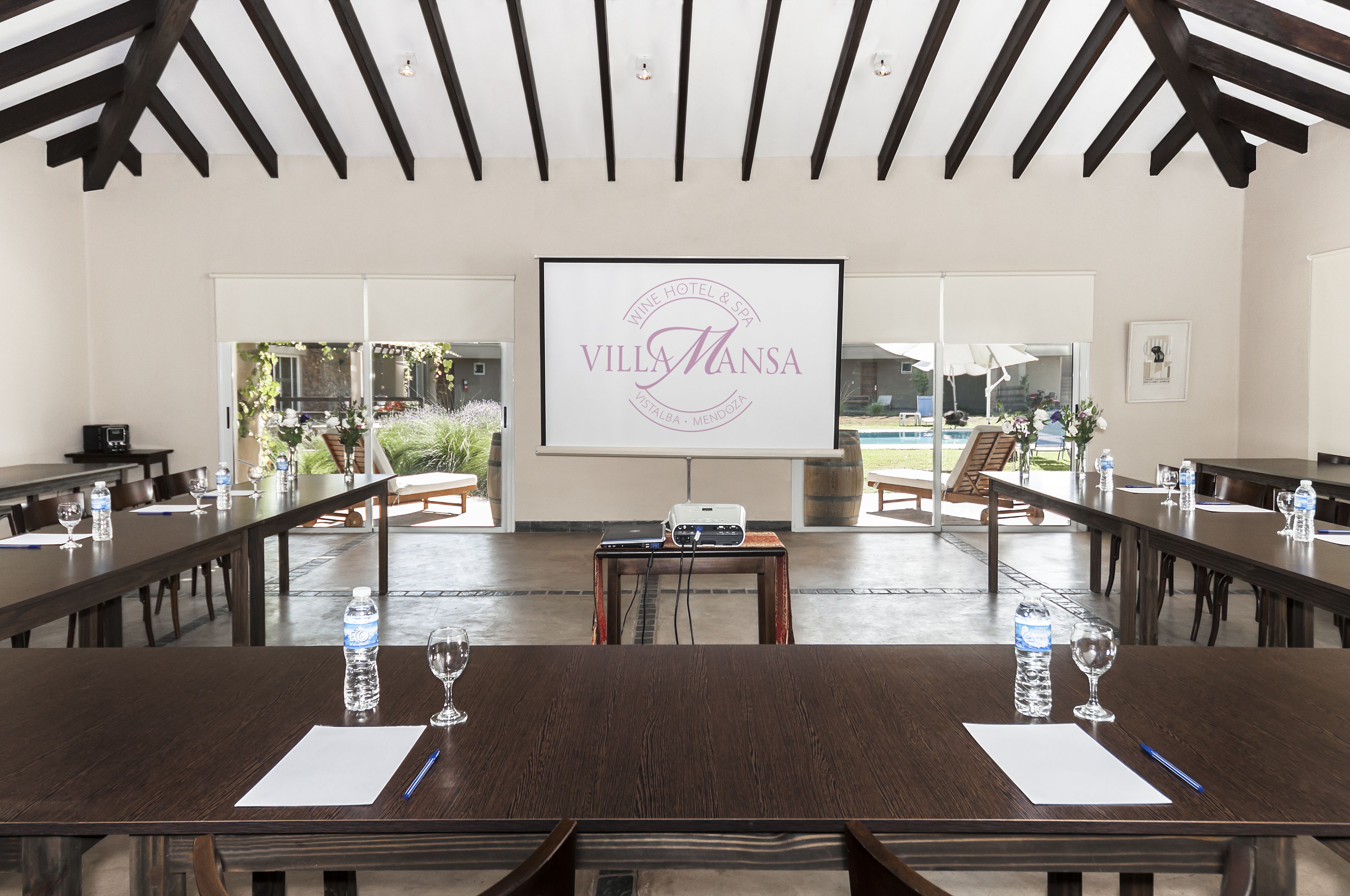 A conference room with a large screen displaying "Villa Mansa" branding, wooden tables with water bottles and glasses, and a pool visible through the glass doors.