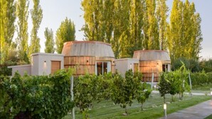 A row of rustic, cylindrical buildings with wooden exteriors, surrounded by lush greenery and trees. - Villa Mansa Wine Hotel (Luján de Cuyo)