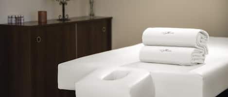 A neatly arranged bathroom with a white towel and a white bath mat on a counter, a wooden cabinet with candles, and a vase with flowers in the background.