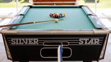 Een pooltafel met een keu en biljartballen, een tafeltennistafel met batjes, en uitzicht op terrasmeubels en groen.