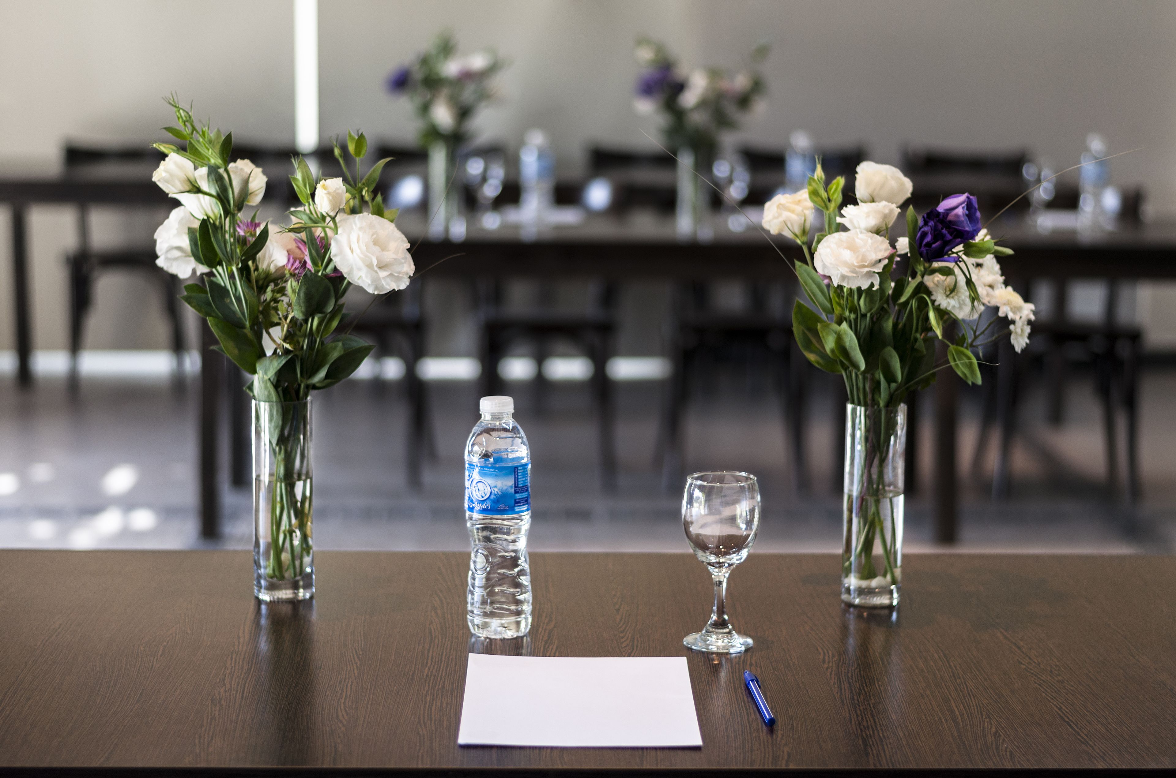 Una mesa de conferencias con jarrones de flores, una botella de agua y una copa de vino.