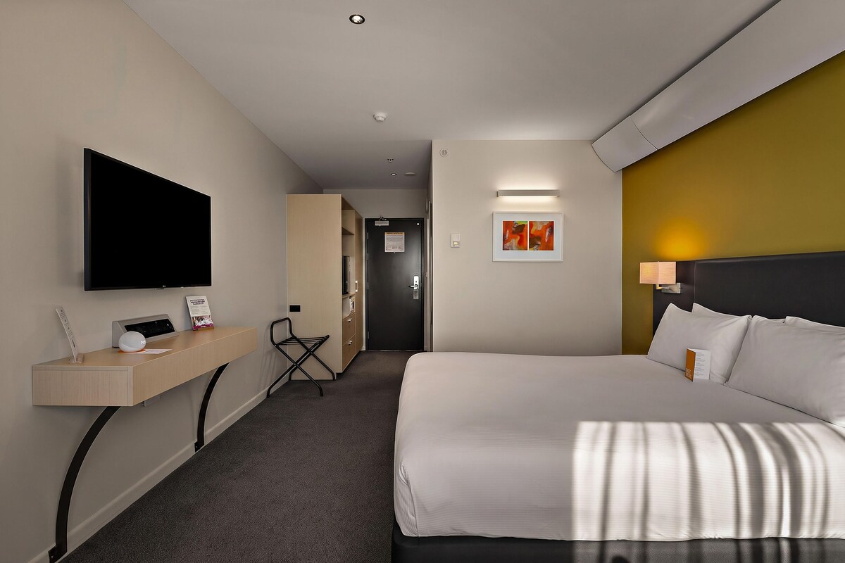Habitación de hotel con cama, escritorio con televisor de pantalla plana, silla, puerta y una obra de arte colgada en la pared.