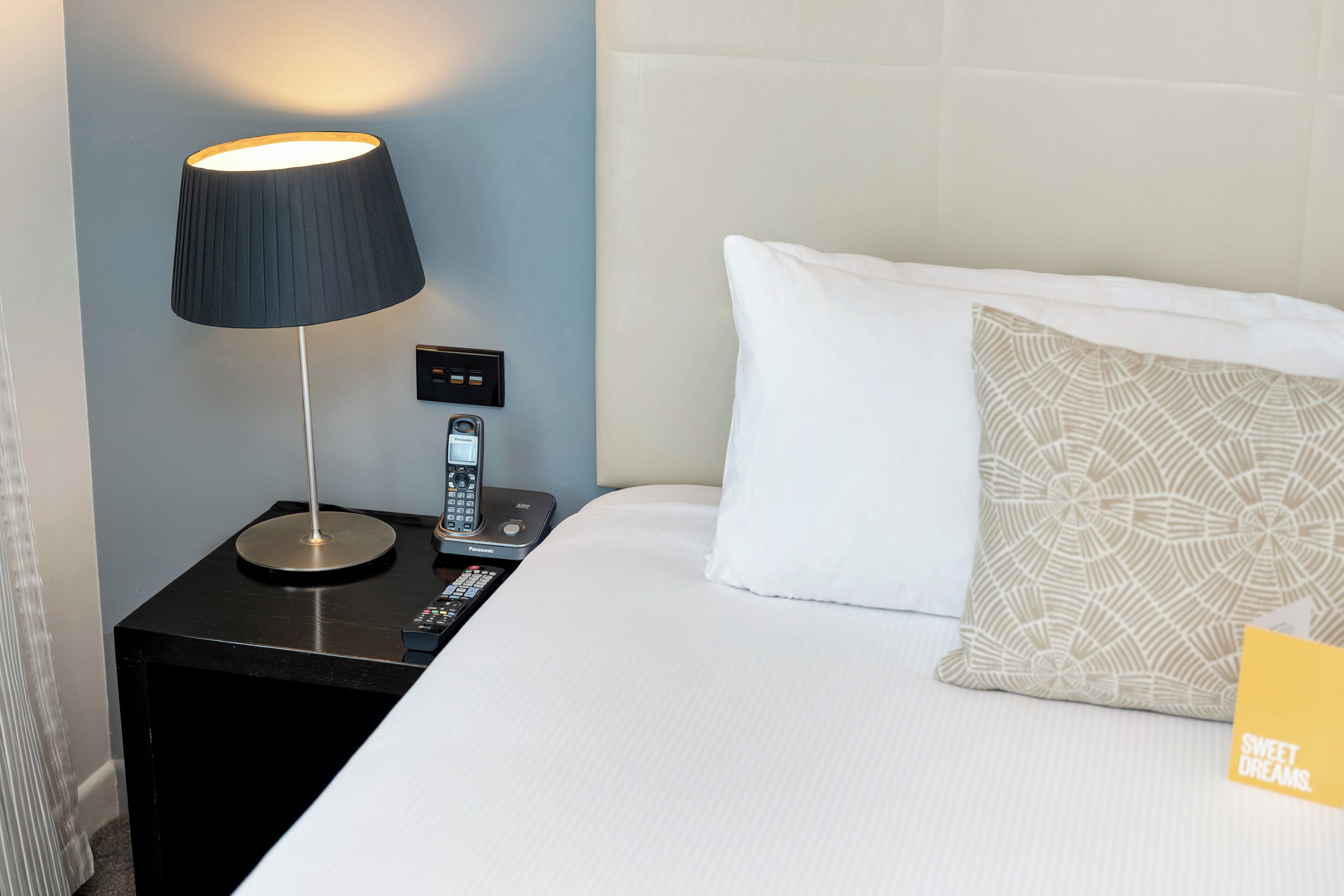 A neatly made bed with a white pillow and a patterned pillow, a black nightstand with a lamp, a remote control, and a telephone.