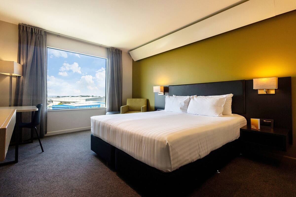 A hotel room with a large bed, a desk, and a chair. There is a window with a view of a building and a sky with clouds.