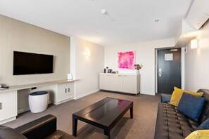 A modern living room with a flat-screen TV, a sofa with cushions, a coffee table, and a kitchen area with cabinets and a countertop.