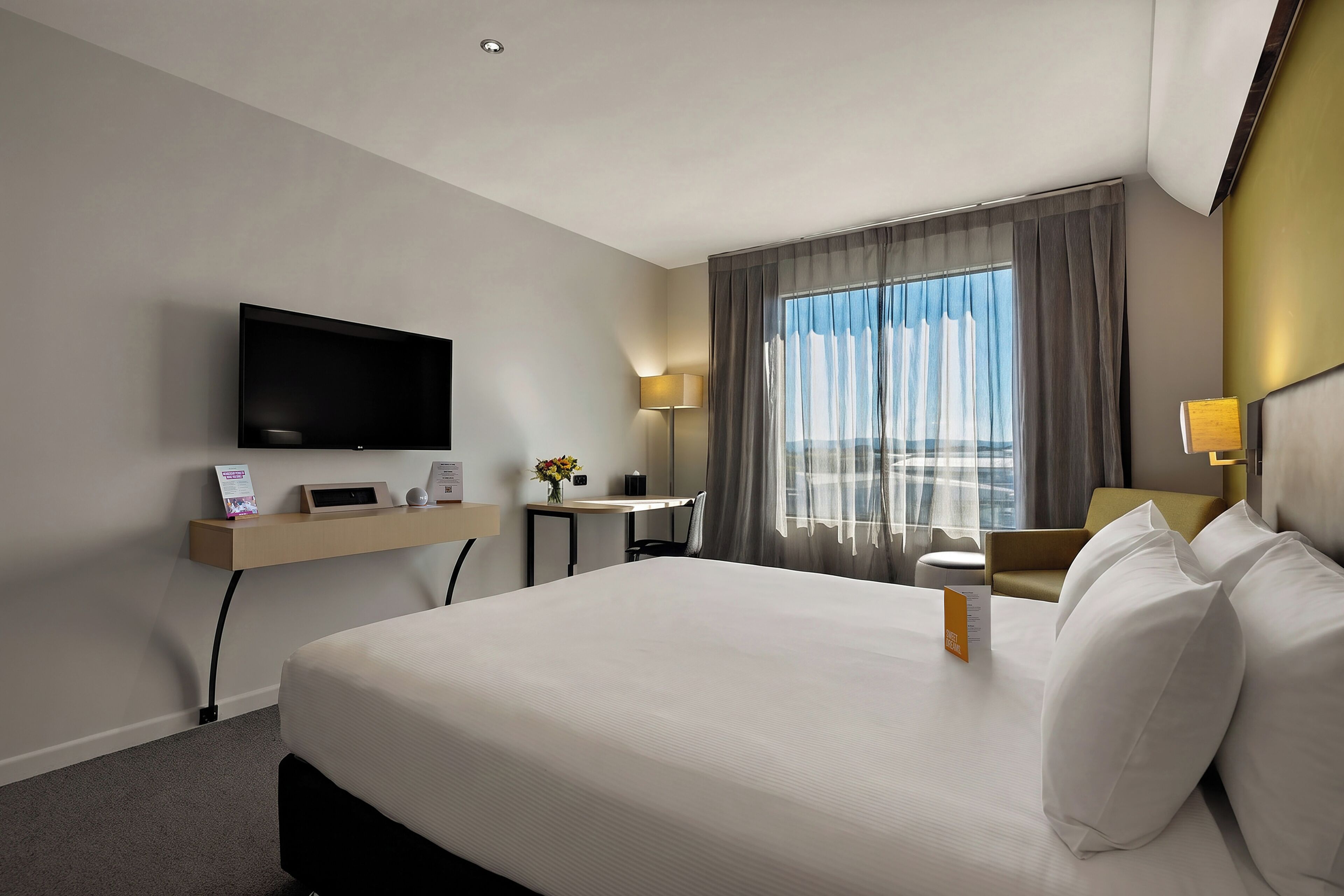 Habitación de hotel con una cama grande, un televisor de pantalla plana montado en la pared, un escritorio con lámpara y una ventana con cortinas.