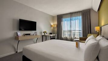 Habitación de hotel con una cama grande, un televisor de pantalla plana montado en la pared, un escritorio con lámpara y una ventana con cortinas.