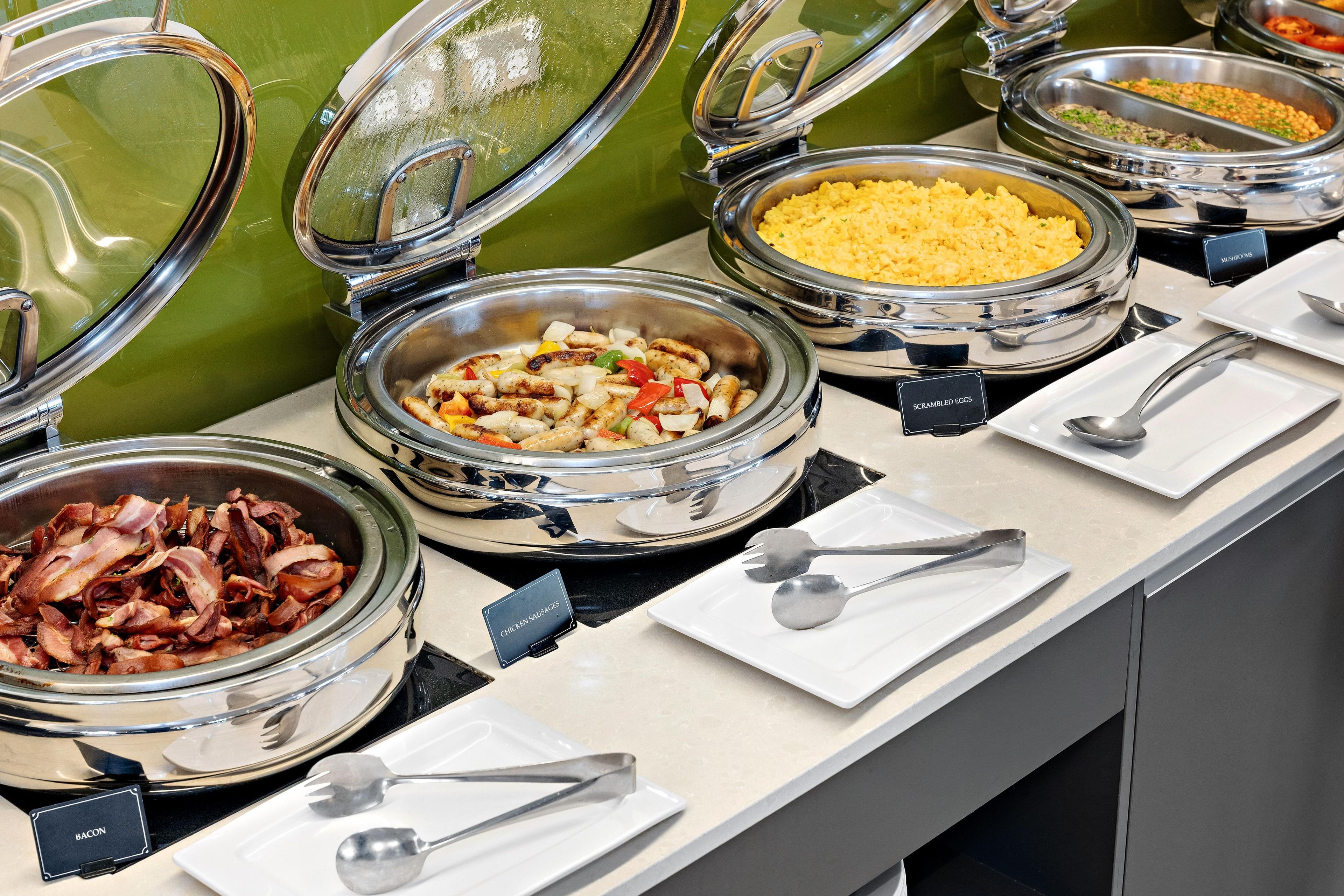 A buffet with various food options including scrambled eggs, chicken sausages, and bacon.