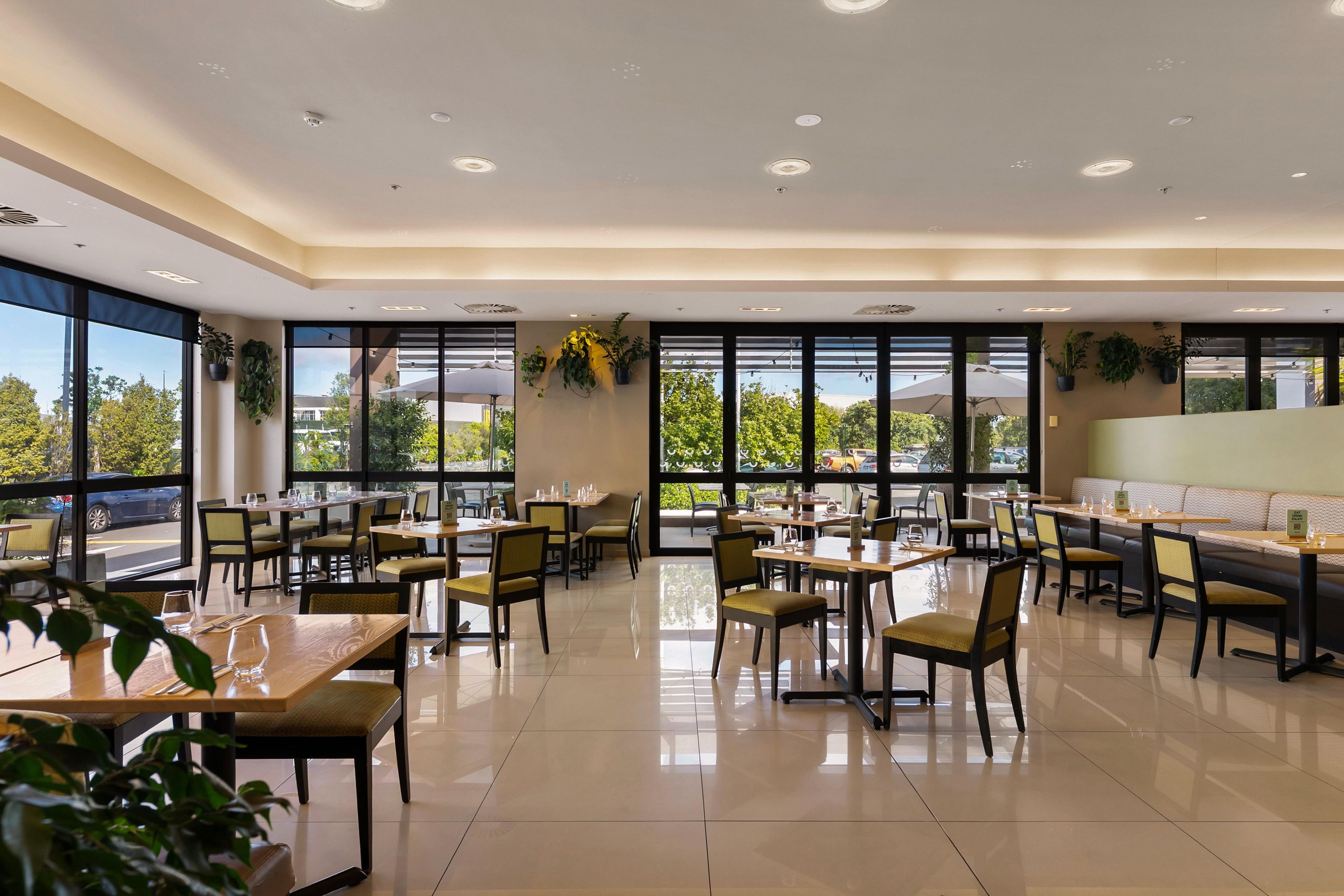 A spacious dining area with multiple tables and chairs, large windows offering a view of the outdoors, and ceiling lights.