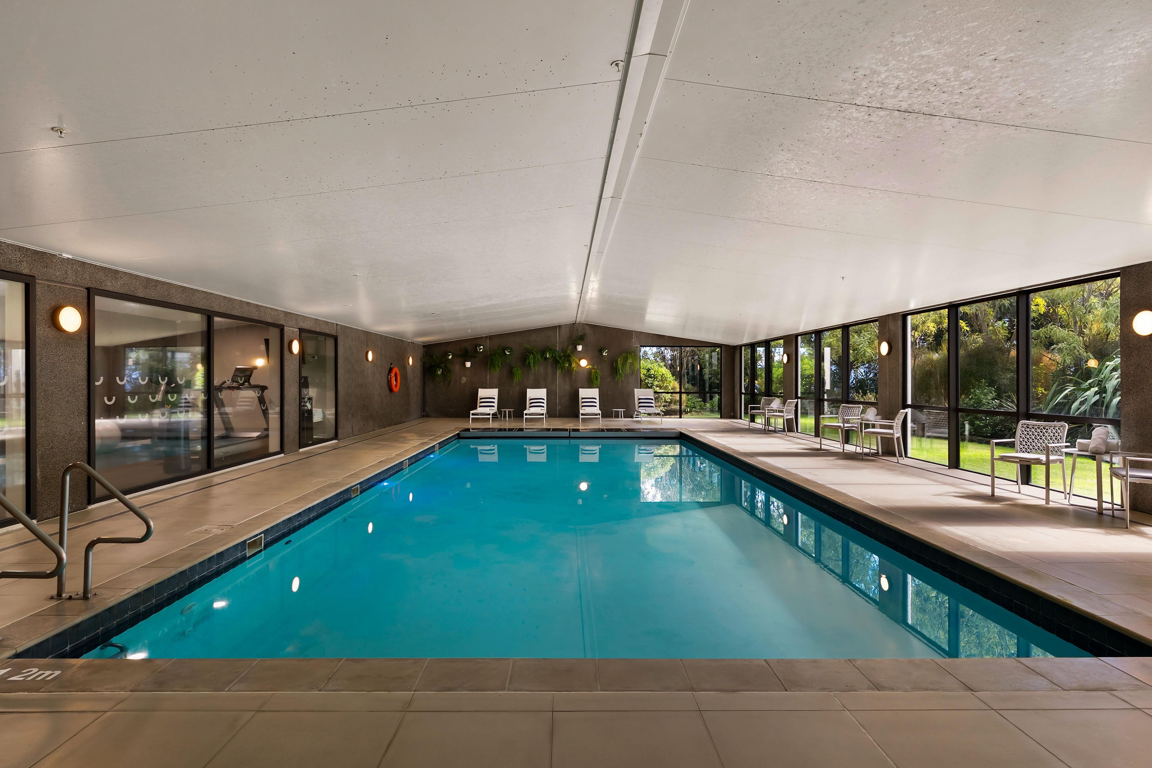 An indoor swimming pool with clear blue water, surrounded by lounge chairs and a view of greenery outside.