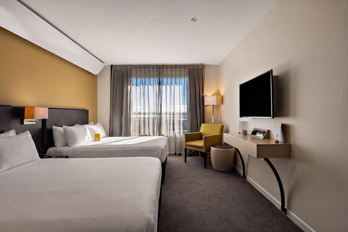 A hotel room with two beds, a desk with a TV, a chair, and a window with curtains.