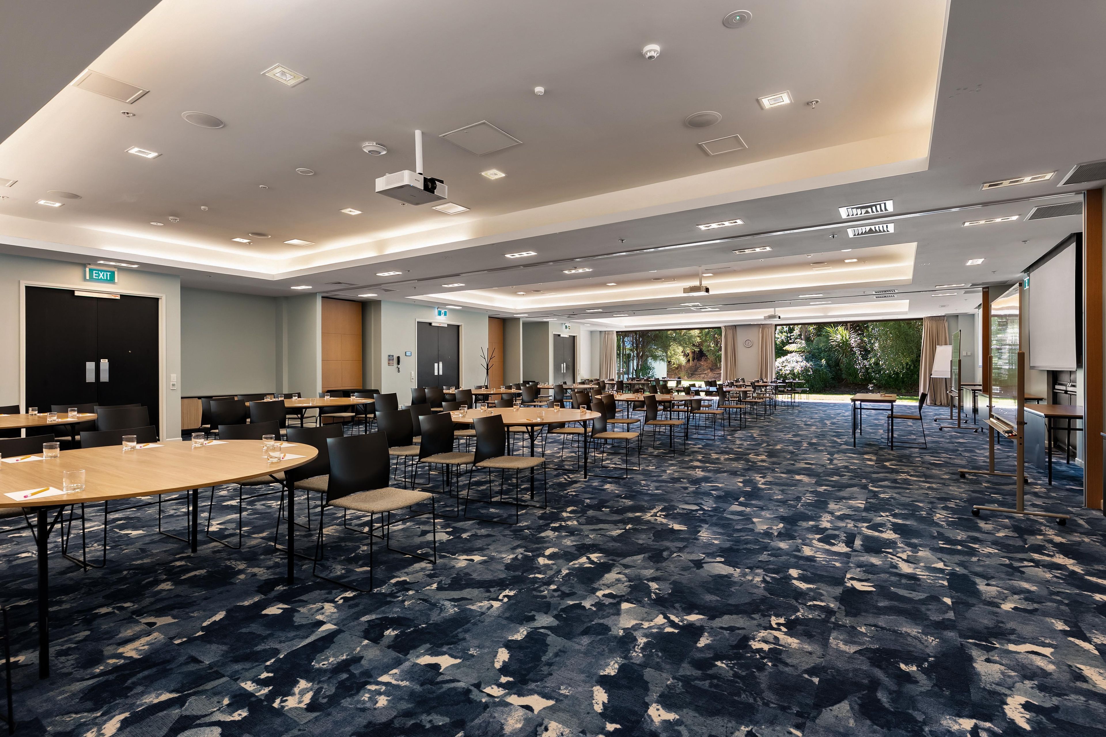 A hotel conference room with multiple tables and chairs, a patterned carpet, and a view of greenery outside.