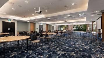 A hotel conference room with multiple tables and chairs, a patterned carpet, and a view of greenery outside.