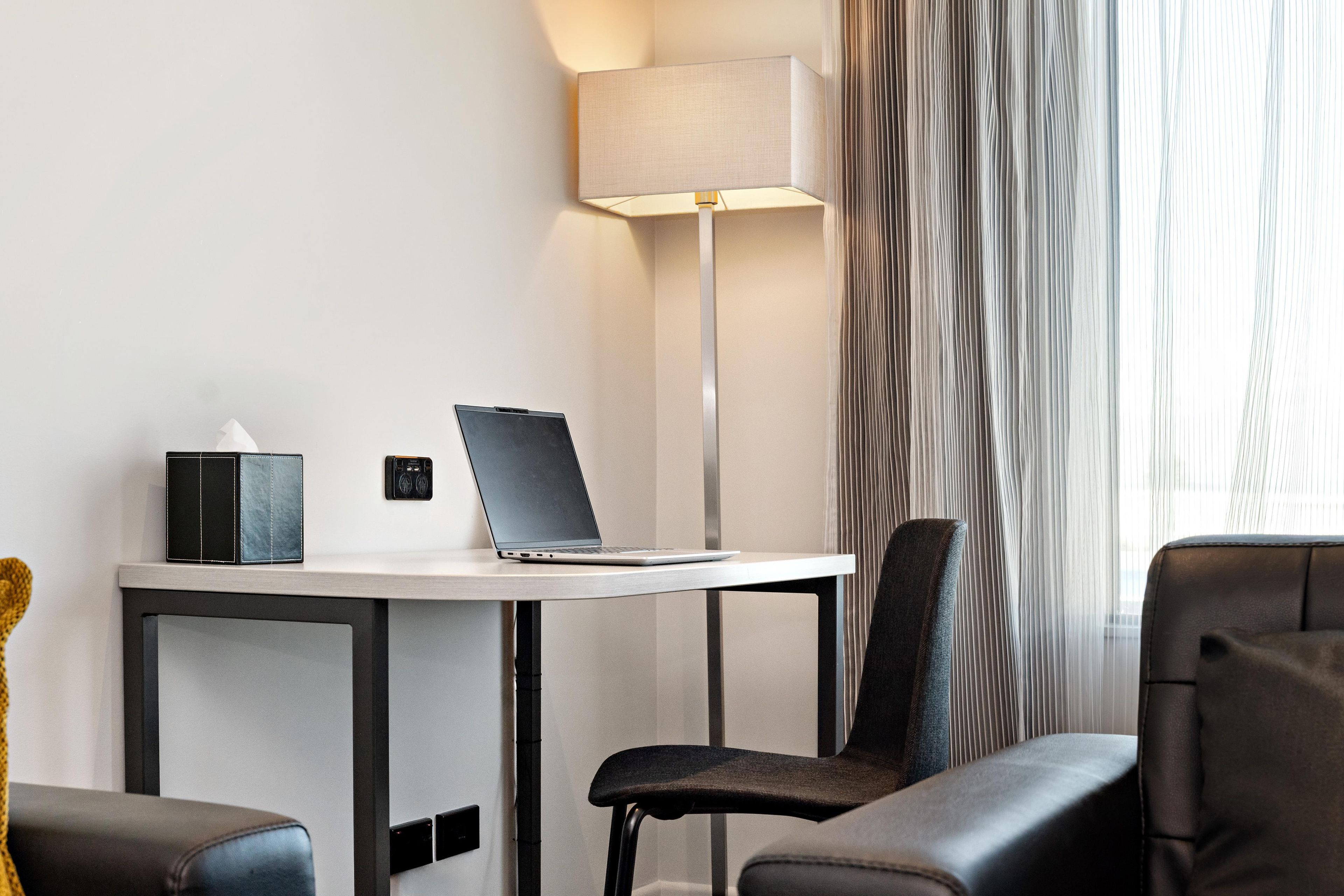 A hotel room with a desk, laptop, lamp, and two chairs.