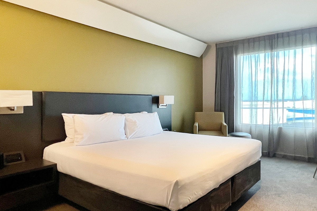 Een hotelkamer met een groot bed, een stoel, een nachtkastje en een raam met gordijnen.