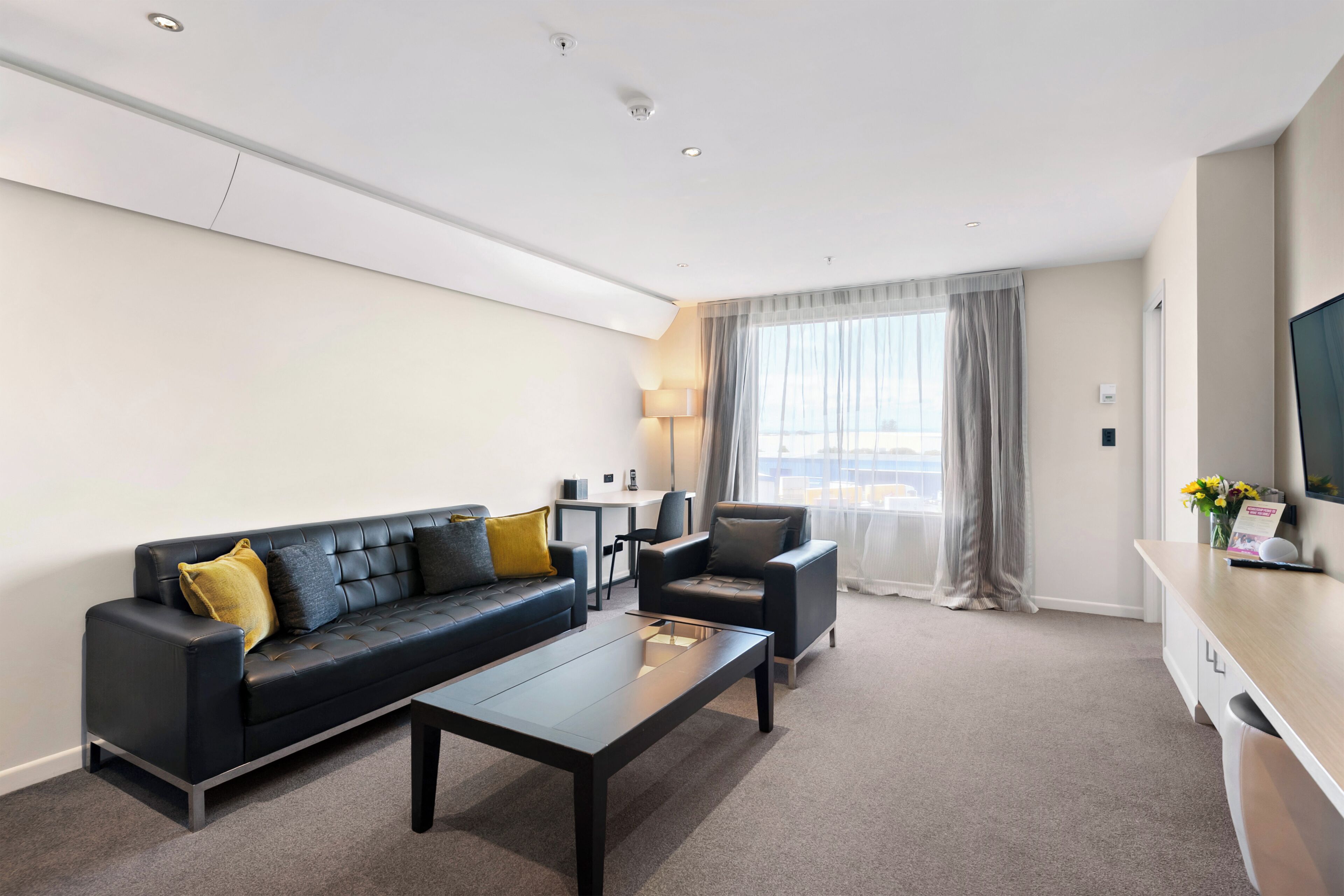 A modern hotel room with a black leather sofa, a small table, a chair, and a large window with curtains.