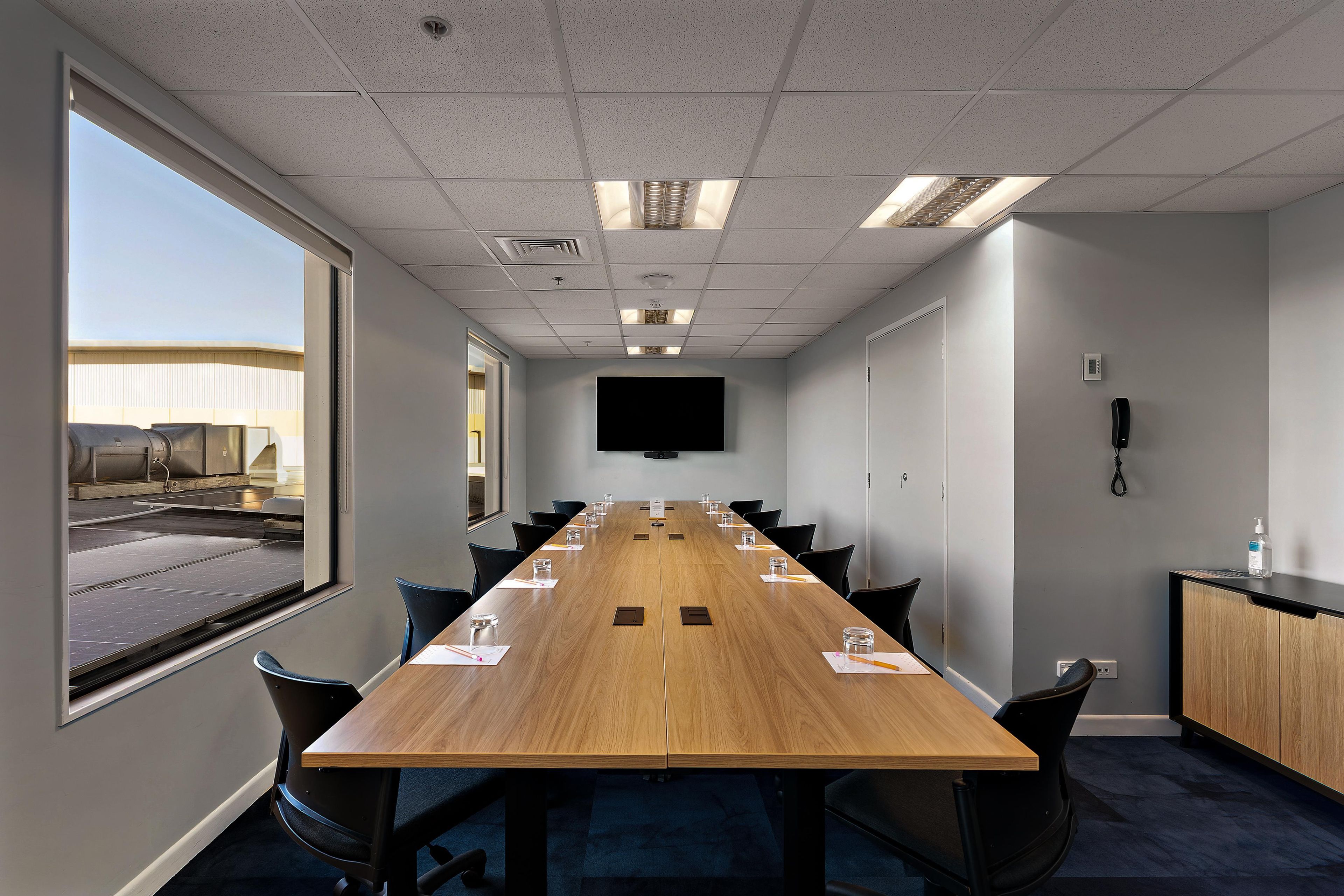A conference room with a long table, chairs, a flat-screen TV, and a window with a view of a building.