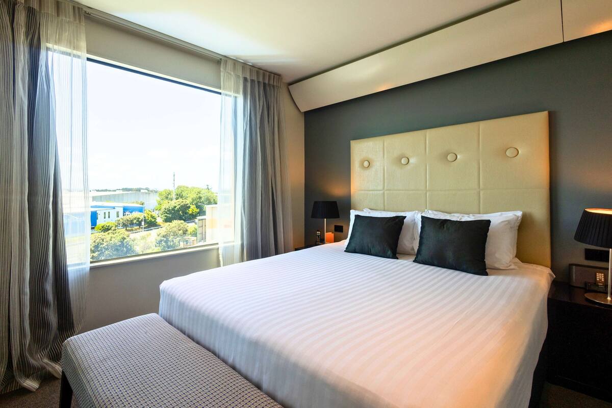 A hotel room with a large bed, a bedside table with a lamp, and a view of buildings and trees through the window.