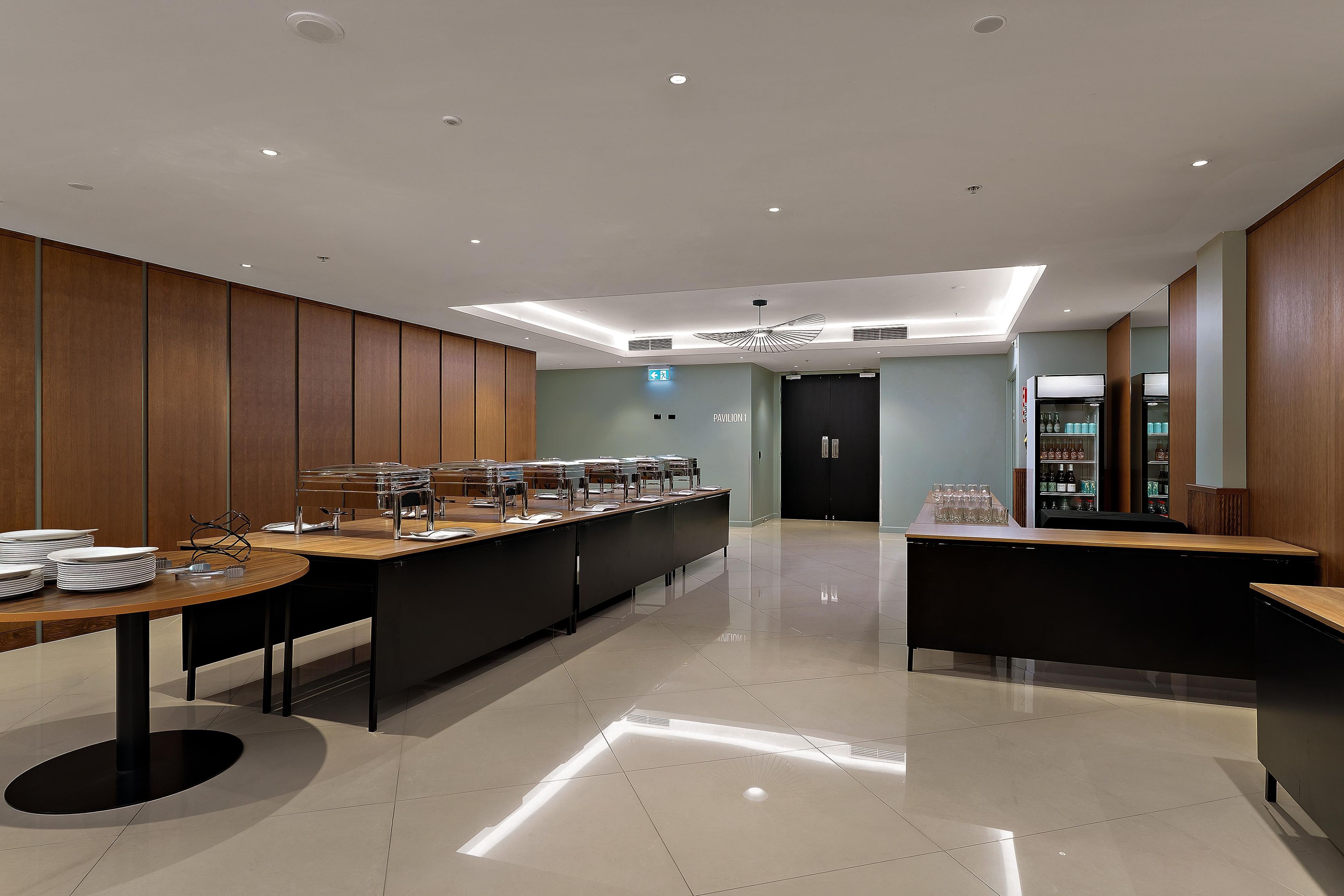 A modern, well-lit room with a long counter for food service, a door labeled 'Pavilion 1,' and a refrigerator stocked with beverages.