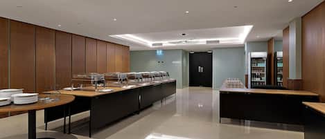 A modern, well-lit room with a long counter for food service, a door labeled 'Pavilion 1,' and a refrigerator stocked with beverages.