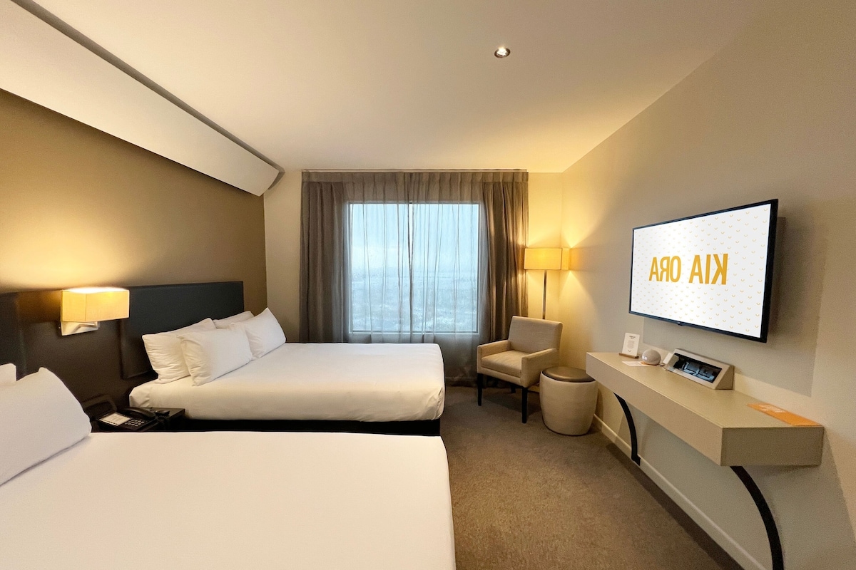 Een hotelkamer met twee bedden, een aan de muur bevestigde televisie, een bureau met een lamp en uitzicht op de omgeving.