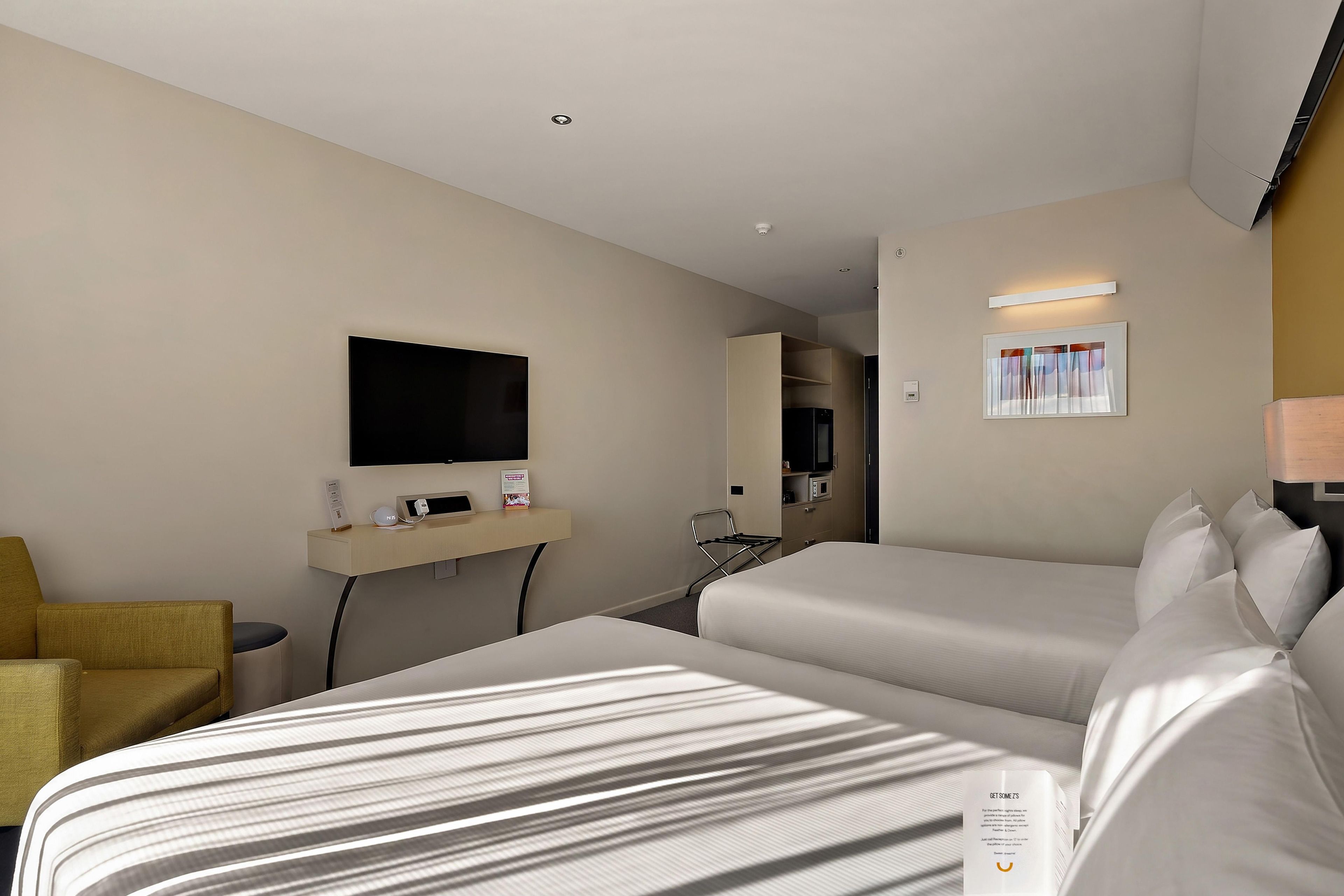 A hotel room with two beds, a television, a desk with a computer, a mirror, and a window with curtains.