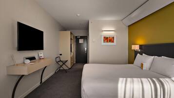 A hotel room with a bed, a desk with a flat-screen TV, a chair, a door, and a wall-mounted artwork.